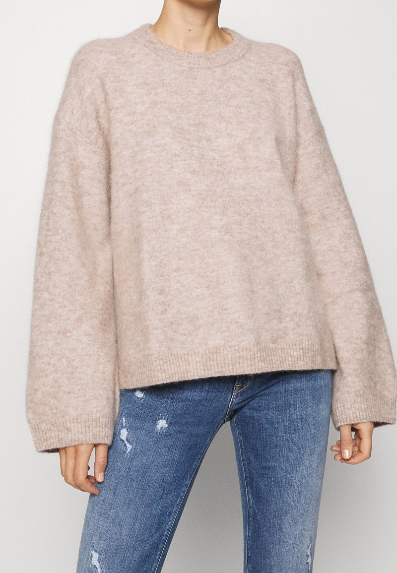 Personne portant un pull en laine beige surdimensionné à manches longues, associé à un jean bleu déchiré, sur fond uni.