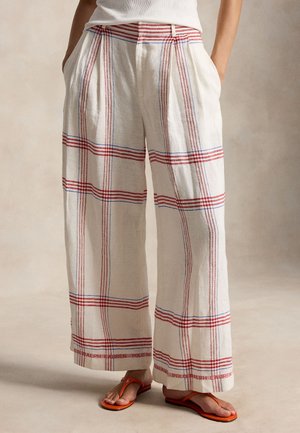 Personne portant un pantalon blanc ample avec des rayures verticales et horizontales rouges et bleues et des sandales orange, les mains dans les poches.