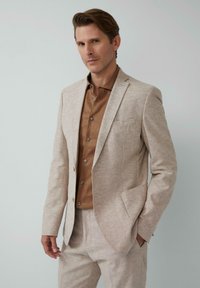 Beige linnenmix pak met een getailleerde pasvorm, een enkele knoop blazer en bijpassende broek, gecombineerd met een bruine overhemd met knopen.