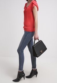 Rote Satinbluse mit kurzen, flatternden Ärmeln, kombiniert mit blauen, eng anliegenden Jeans. Die schwarze Handtasche hat eine strukturierte Oberfläche, und die Stiefeletten besitzen eine spitze Zehenform.