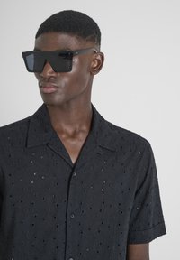 Camisa de manga corta negra con un diseño floral recortado, combinada con gafas de sol negras de gran tamaño con montura angular.