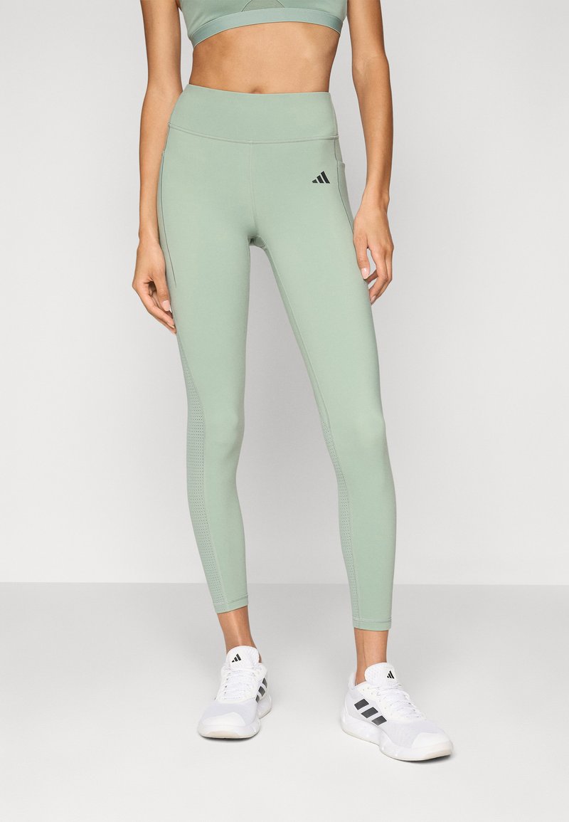 Világoszöld leggings sima textúrával, magas derékkal, oldalsó varrás részletezéssel és egy kis fekete logóval a bal combon. Fehér sportcipőkkel stylingolva.