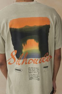 Olive green T-shirt featuring a large graphic print with orange, black, and green hues, text reading "Silhouettes" and intricate patterns.