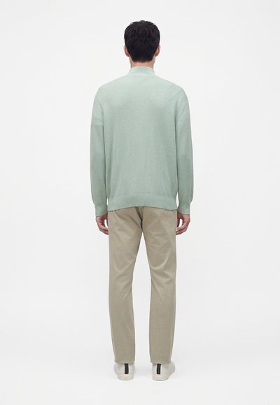 Homme debout de dos, portant un pull vert clair, un pantalon beige et des baskets blanches avec des bandes noires sur un fond blanc uni.