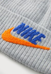 Pilkos juostos raštuotos megztos skrybėlės su mėlynu ir oranžiniu siuvinėjimu Nike logotipo. Medžiaga yra minkšta, matomi megzti raštai visame paviršiuje.