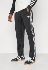 Μαύρο αθλητικό παντελόνι με λευκές ρίγες στα πλαϊνά, με μικρό λευκό λογότυπο και κίτρινες λεπτομέρειες, κατασκευασμένο από μαλακό ύφασμα με τσέπες.