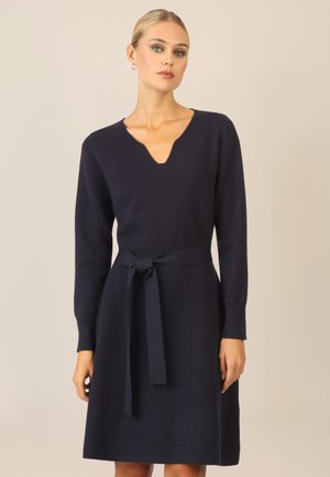 Navy-blaues Strickkleid mit V-Ausschnitt, langen Ärmeln und gebundenem Tailleneinsatz. Der Stoff wirkt weich mit einer glatten Textur und hat ein knielanges Design.