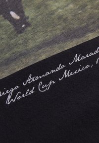Sort T-shirt med et vintage print af en fodboldscene, med "Diego Armando Maradona World Cup Mexico" i hvidt kursivskrift. Ensartet tekstur.