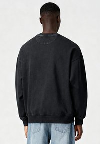 Personne à la peau foncée et aux cheveux courts, portant un sweat-shirt noir ample et un jean bleu clair, debout de dos face au spectateur sur un fond blanc.