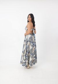 Robe maxi en tissu léger, arborant un motif de feuilles en bleu et beige. Dos ouvert avec des liens. Jupe évasée et chaussures à talons hauts visibles.