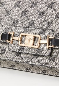 Primo piano di una borsa a spalla con tessuto testurizzato bianco e nero e una chiusura rettangolare dorata con un piccolo emblema floreale.