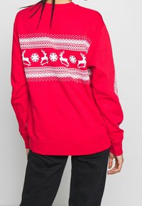 Sweat-shirt rouge avec un motif de rennes blancs et flocons de neige sur le dos, doté d'un col rond et de poignets côtelés. Texture de tissu doux.