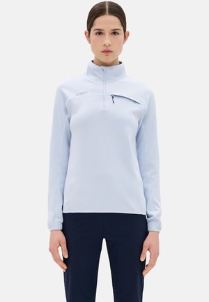 Jong persoon met donker haar draagt een lichtblauwe trui met kwartritssluiting en donkere broek, staand tegen een effen witte achtergrond.