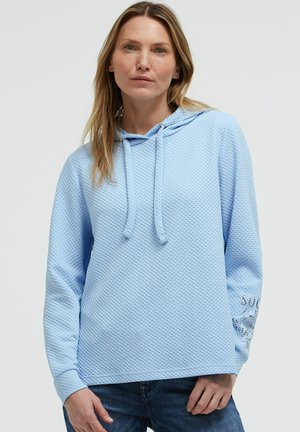 Frau trägt hellblauen strukturierten Hoodie mit Kordelzug und Jeans, steht vor einfarbigem Hintergrund.