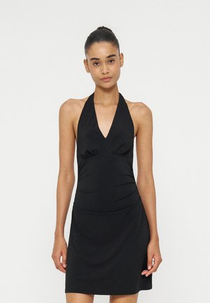 Jeune femme avec les cheveux attachés en chignon, portant une robe noire sans manches à col halter, debout devant un fond blanc uni.