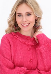 Cardigan lavorato a maglia di un vivace rosa con colletto arricciato, maniche texturizzate e bottoni rotondi rosa. Gli orecchini abbinati sono decorativi.