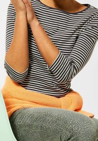 Randig svart och vit långärmad skjorta, orange stickad tröja knuten runt midjan och strukturerade grå byxor, synliga från midjan och uppåt.