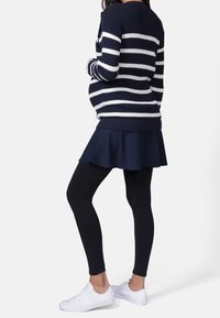 Těhotná osoba ve svetru s námořnickými a bílými pruhy, námořnické sukni, černých legínách a bílých teniskách stojící bokem na bílém pozadí.