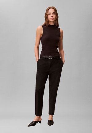 Kvinde iført sort ærmeløs top, sorte bæltebukser og sorte lave hæle, stående med hænderne i lommerne mod en ensfarvet baggrund.