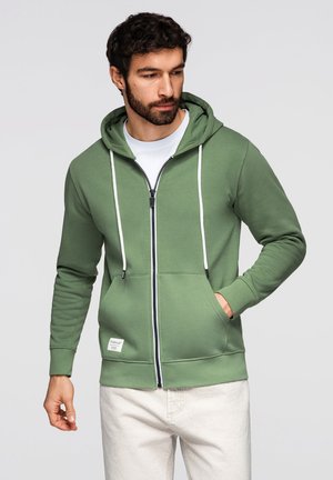 Man met donker haar en baard, gekleed in een groene hoodie met rits en witte trekkoorden, en lichtgekleurde broek, tegen een effen achtergrond.
