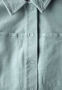 Camicia in corduroy verde chiaro con colletto a punta, due tasche frontali e bottoni rotondi con testo. Il tessuto presenta una texture a coste visibile.