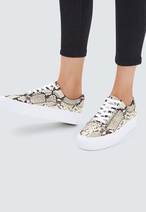 Feet wearing low-cut sneakers with white thick soles and a beige and black snakeskin pattern, paired with black cropped pants.