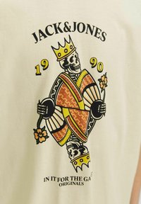 Maglietta in cotone color crema con una grafica di uno scheletro colorato che tiene dei fiori, con una corona e motivi decorativi, e del testo sottostante.