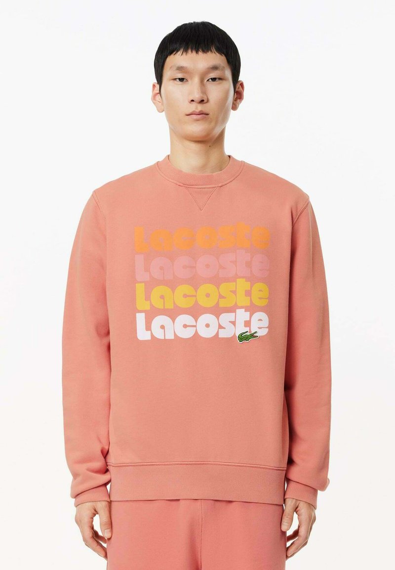 Felpa rosa con scollo rotondo, con il testo "Lacoste" in più colori e un accento di logo coccodrillo verde sul davanti.