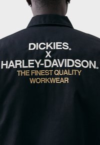 Black workwear jacket with embroidered text featuring "DICKIES x HARLEY-DAVIDSON" in white and "THE FINEST QUALITY WORKWEAR" in gold.