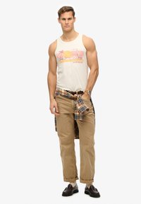 Camiseta sin mangas blanca con un gráfico del sol, pantalones caqui y una camisa de cuadros atada a la cintura. Unos mocasines marrones y un reloj complementan el conjunto.