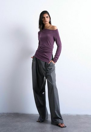 Femme vêtue d'un haut violet à épaules dénudées et d'un pantalon gris à jambes larges, debout les mains dans les poches sur un sol gris contre un mur blanc.