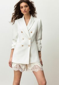 Weißer, doppelreihiger Blazer mit goldfarbenen Knöpfen, langen Ärmeln und tailliertem Schnitt, kombiniert mit cremefarbenen Shorts mit Spitzenbesatz.