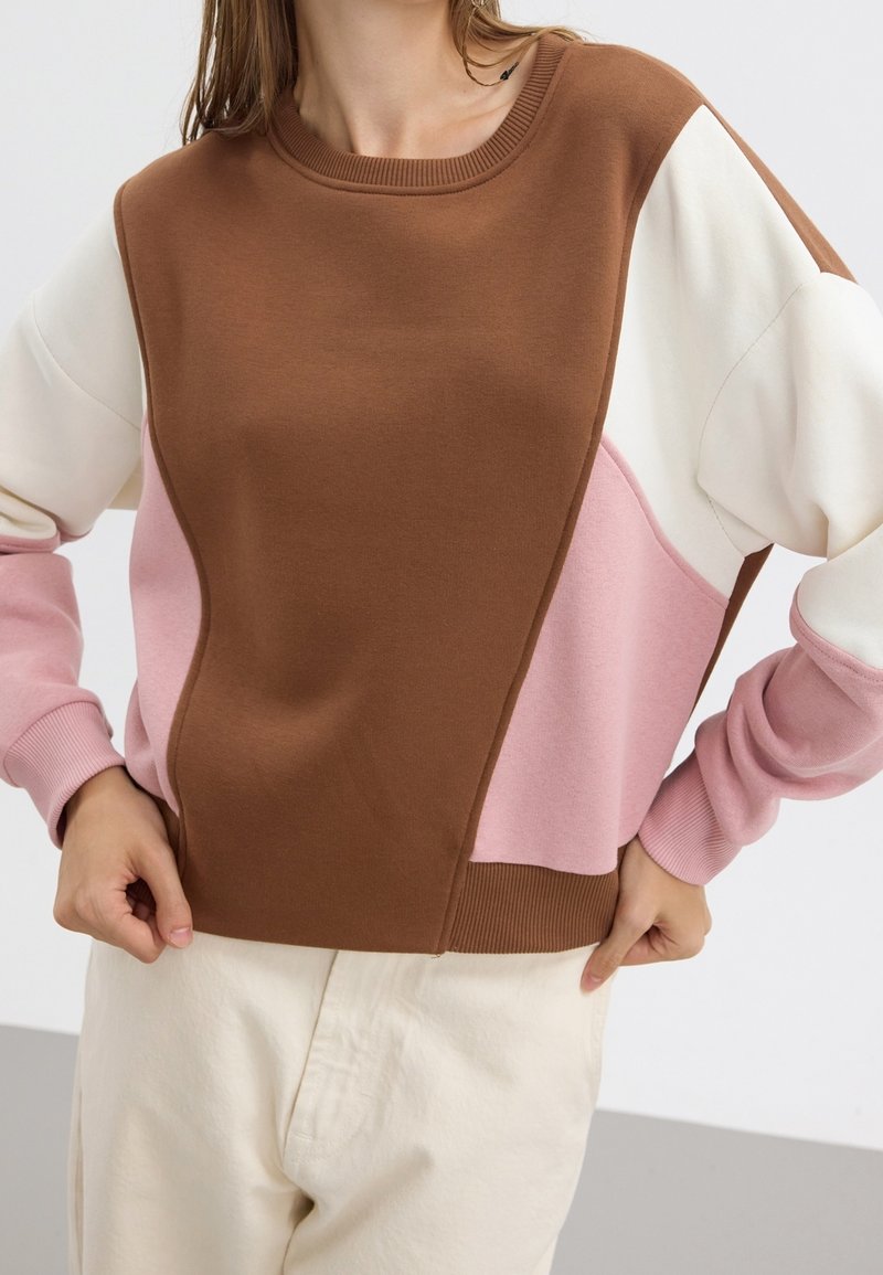 Kobieta ubrana w sweter z blokami kolorów w odcieniach brązu, różu i bieli, w połączeniu z jasnobeżowymi spodniami, stojąca z rękami na biodrach.