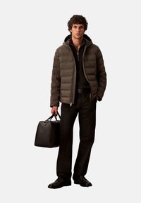 Chaqueta tipo plumas marrón oscuro con capucha, camiseta negra con cremallera, pantalones negros, sosteniendo una bolsa negra con asas y usando zapatos negros.