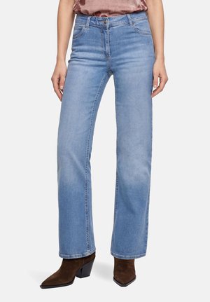 Femme portant un jean droit bleu clair et des bottes à talons en daim marron, debout, les mains détendues le long du corps.