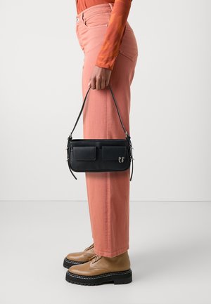 Czarna skórzana torba z dwiema przednimi kieszeniami, trzymana przez osobę w jasnoróżowych szerokich spodniach i beżowych butach na sznurowadła.