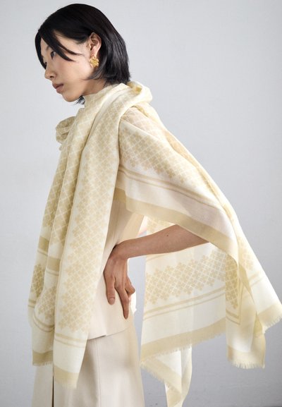 Écharpe beige avec un motif géométrique, drapée sur les épaules. Fabriquée en tissu doux avec des bords frangés, stylisée sur une tenue crème.