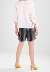 Blouse blanche semi-transparente avec des manches trois-quarts et des accents en lien, associée à des shorts rayés noirs et blancs. Mules à talons jaunes.