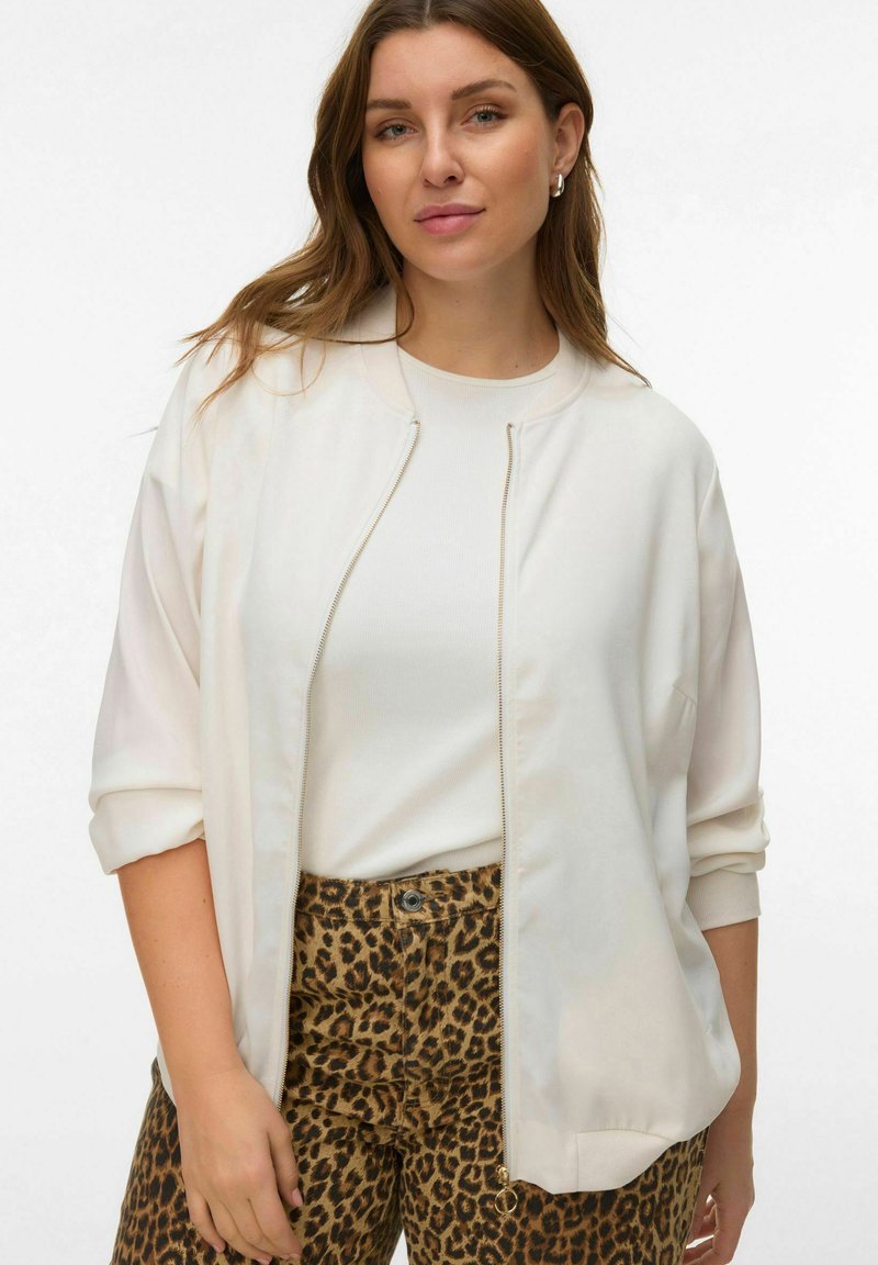 Veste bomber zippée ivoire à épaules tombantes, associée à une chemise crème claire et un pantalon taille haute imprimé léopard marron. Texture lisse.