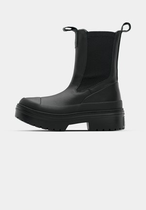 Bottines noires à cheville avec une tige synthétique texturée, des panneaux latéraux élastiques et une semelle épaisse et robuste. Elles sont munies de languettes pour un enfilage facile.