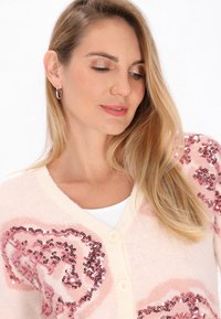 Rosa Strickjacke mit Paillettenherzmuster, cremefarbene Knöpfe, weicher Textur und V-Ausschnitt, getragen über einem weißen Oberteil.