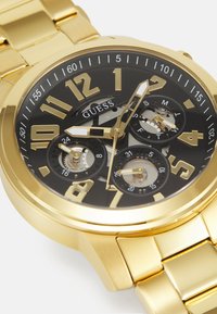 Reloj Guess en tono dorado con una esfera negra, marcadores numéricos prominentes, funciones de cronógrafo y un diseño elegante de brazalete de metal.