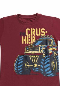 Bordeaux t-shirt met een grafiek van een monstertruck met overmatige blauwe banden, zwarte accenten en gele tekst met de woorden "THE CRUSHER."
