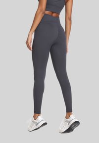 Kobieta w ciemnoszarych legginsach o wysokim stanie i białych butach sportowych, stojąca na jednolitym, jasnoszarym tle.