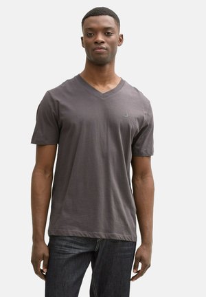 Camiseta de algodón gris con cuello en V y mangas cortas, con un sutil logo en el pecho, combinada con jeans de denim oscuros. Textura suave y diseño casual.