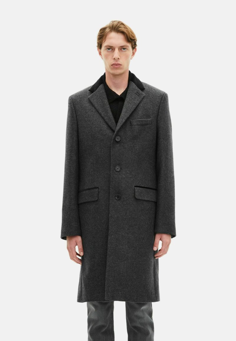 Grå hørflet uldfrakke med tre knapper, slå-om krave og to frontlommer. Båret over en sort skjorte og grå bukser.