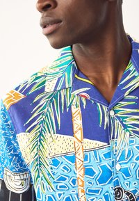 Chemise à manches courtes en bleu vif avec des motifs tropicaux de palmiers et géométriques, dotée d'un col camp et de boutons, fabriquée en tissu léger.