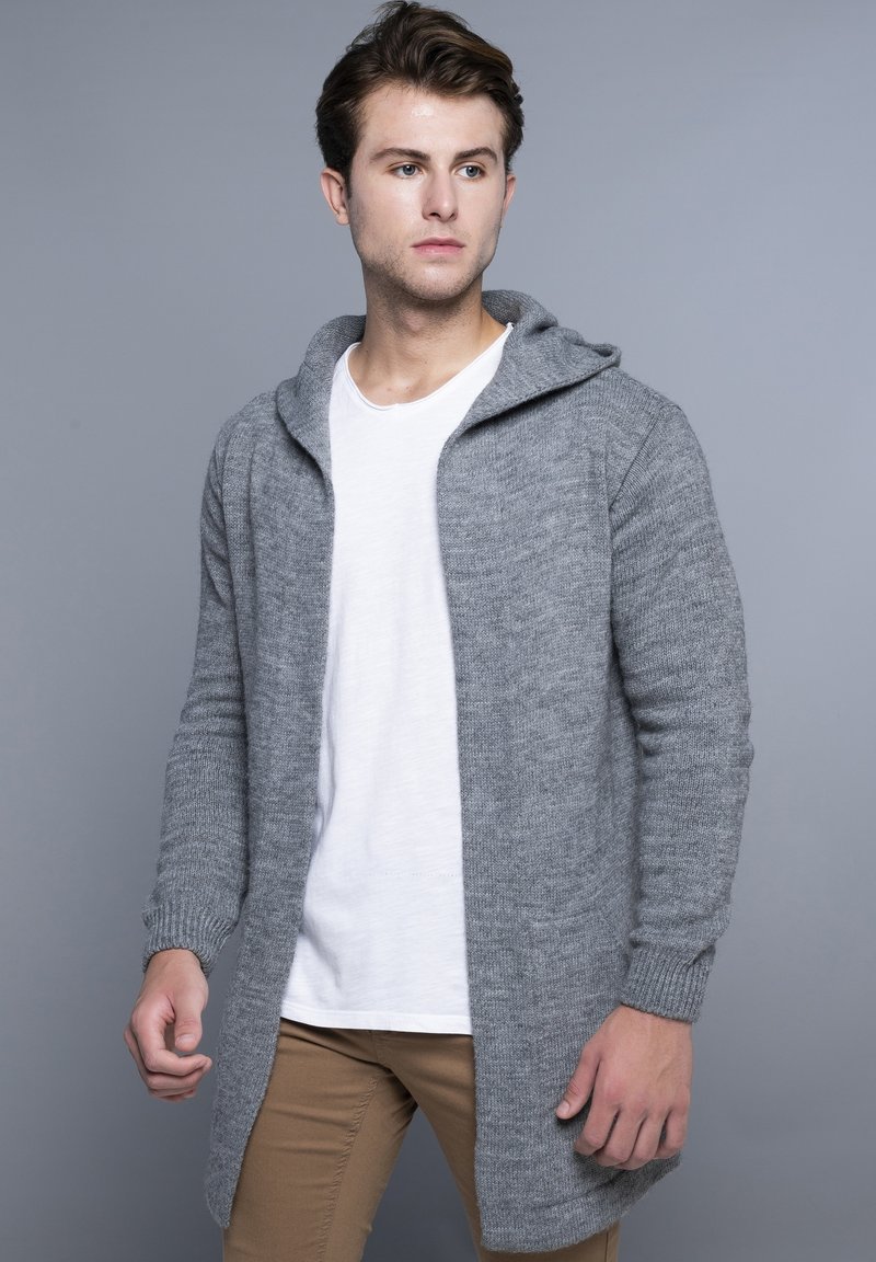 Lysegrå strikket cardigan med hætte, med frontåbning og ribbede ærmer. Bærer den over en hvid T-shirt og brune bukser.