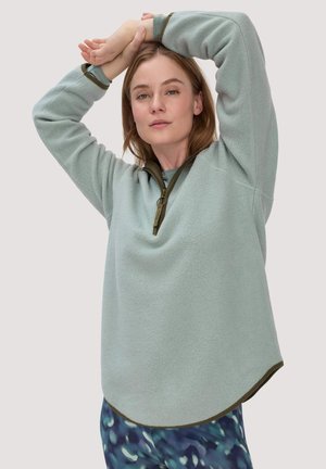 Junge Frau, die die Arme über dem Kopf ausstreckt und einen hellgrünen Fleecepullover mit halbem Reißverschluss sowie blaue, gemusterte Hosen vor einfarbigem Hintergrund trägt.