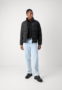 Svart boblejakke med høy krage, glidelås og side lommer, kombinert med en svart polo, lyseblå jeans og svarte sko.